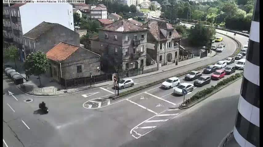 A motorcycle drives through a street intersection near a cluster of buildings on a sunny day, with several cars parked and driving along a nearby road curving around a hillside.
