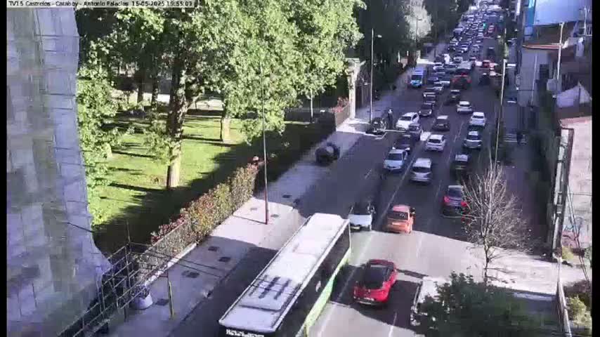 A long line of cars is driving down a city street on a sunny day, beside a grassy park and a tall building, with a bus driving in the opposite direction.