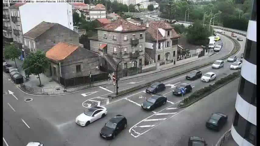 An overcast day shows a street intersection with several cars navigating a roundabout and a two-story building with a gabled roof in the background.