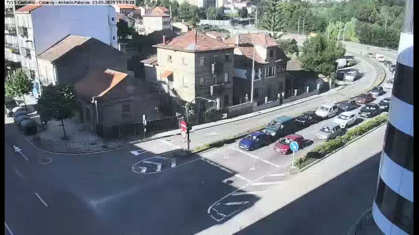 A sunny day shows a street intersection with several cars stopped and moving, near some buildings and a hill with greenery.