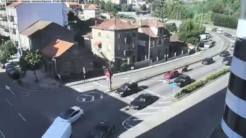 A sunny day reveals a street intersection with several cars driving, a few buildings, and some greenery.