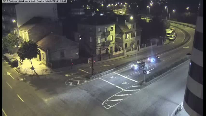 At night, a few cars drive along a multi-lane road that curves around a building; several parked cars and buildings are visible in the surrounding area.