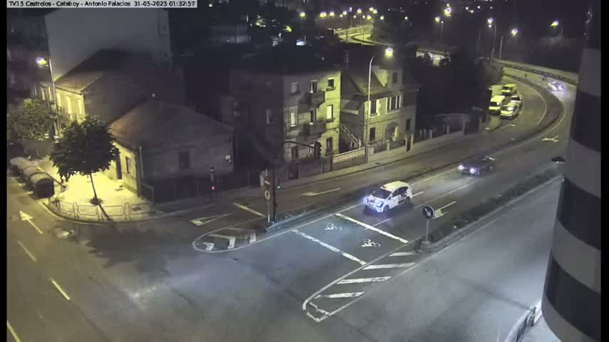 An aerial nighttime view of a street intersection with several cars, and buildings lit by streetlights.