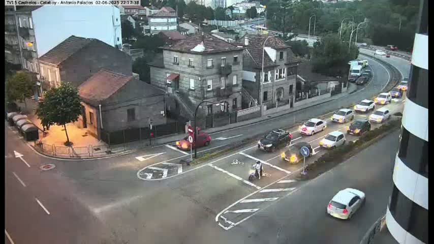 Several cars are driving on a street intersection at dusk, near buildings.