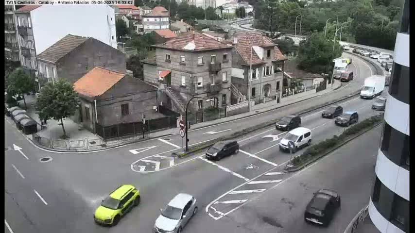 A high-angle view shows a city intersection with several cars at a stop sign and a road curving around a hill with more cars and buildings in view, under partly cloudy skies.
