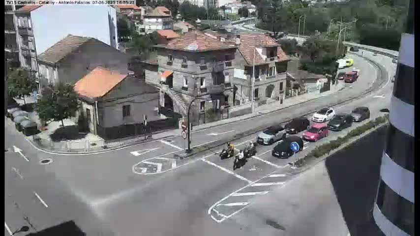 A street scene on a sunny day shows several cars parked and driving on a road that intersects with another road, with several buildings visible, including a multi-story building and a few smaller houses, and several motorcyclists are at an intersection.