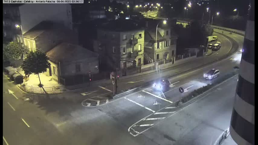 A nighttime street scene shows a few cars at an intersection near buildings under streetlights.