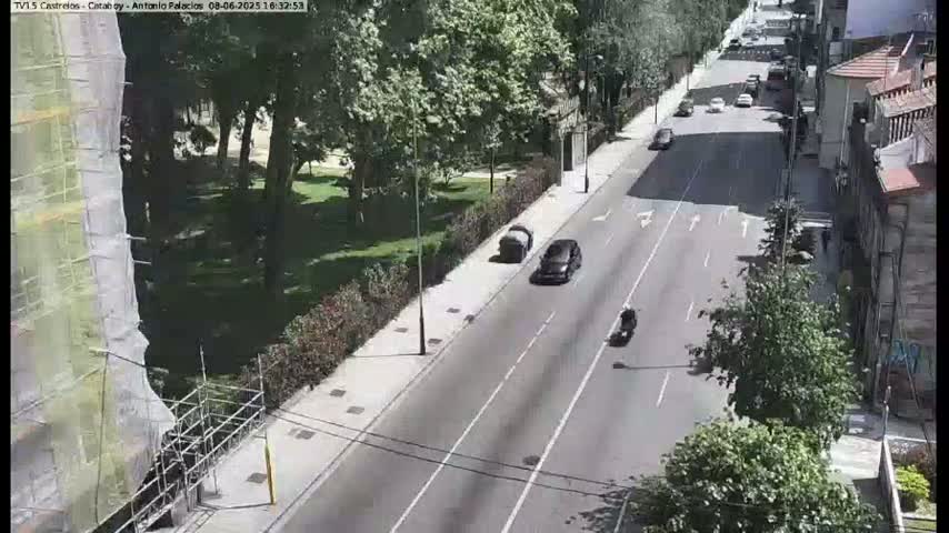 A sunny day shows a city street with several cars and a motorcycle driving, next to a park with lush green trees and a building under construction.