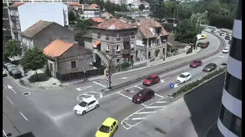 A sunny day shows several cars navigating a multi-lane roadway and intersection that winds around buildings in a residential area.