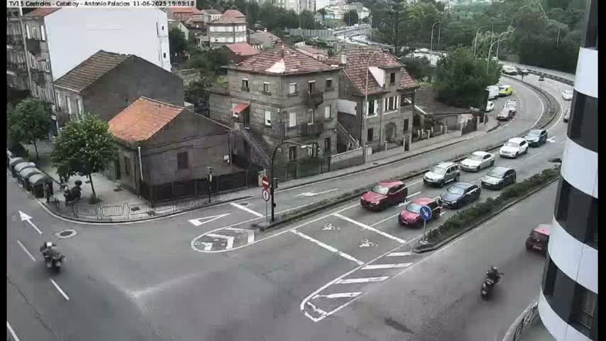 A street intersection with several cars and motorcycles, surrounded by buildings and trees under an overcast sky.
