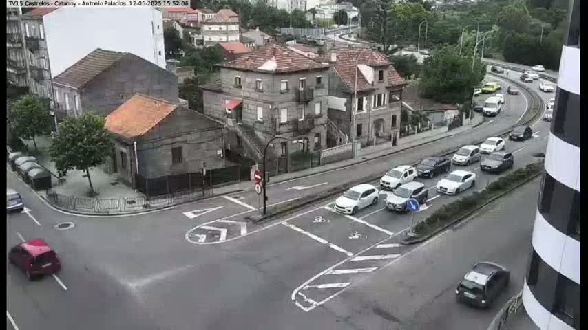 Several cars are driving on a multi-lane road that intersects with a smaller street in front of some buildings, on an overcast day.