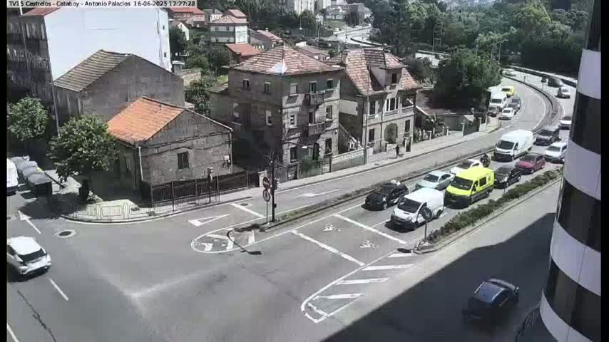 A sunny day reveals a street intersection with several cars and buildings, including a multi-story residential building, and a road curving uphill with moderate traffic congestion.