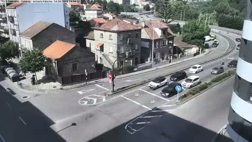 A sunny day shows several cars driving around a street corner near multiple buildings.