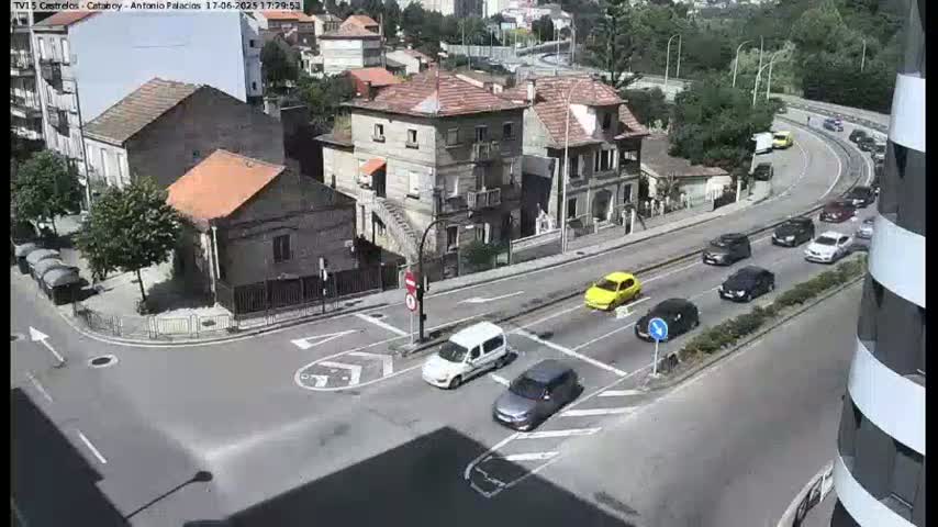 A sunny day shows a street intersection with several cars, a white van, and buildings, including a multi-story building with a red tile roof.