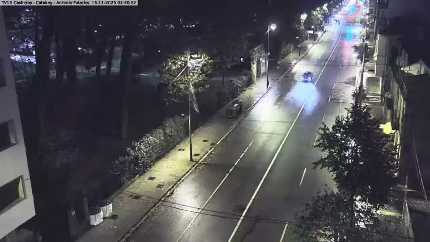 A nocturnal, high-angle view captures a wet city street where a car with bright headlights leaves a luminous motion blur streak, driving past streetlights, buildings, and a park, suggesting recent rain.