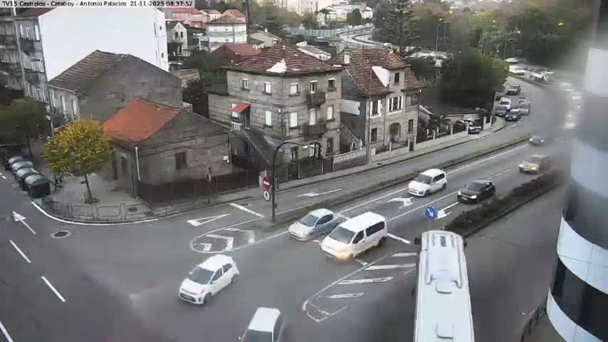 An overhead view captures vehicles navigating a busy urban intersection bordered by residential buildings and trees, all under an overcast sky.