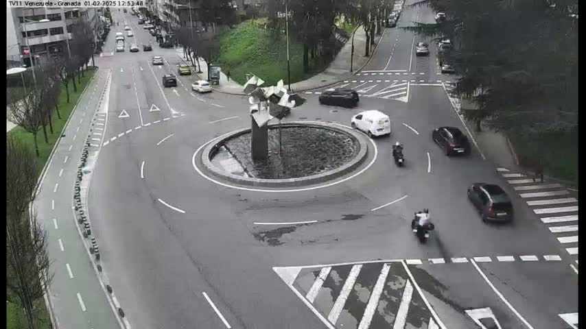 A roundabout with several cars and motorcycles driving around it on a gray day.