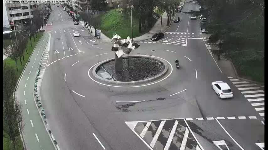 A roundabout with a modern sculpture in the center is surrounded by roadways with several cars and a motorcycle, under a cloudy sky.