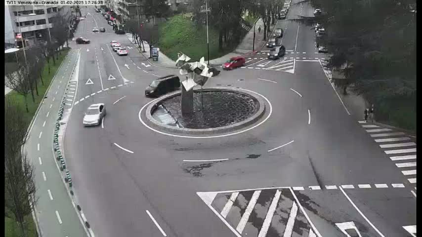 A roundabout with a modern sculpture in the center is surrounded by several cars on a mostly overcast day.