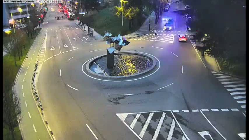 A circular traffic island with a modern sculpture in the center is surrounded by roads with cars and a pedestrian crossing under a dimly lit, nighttime sky.