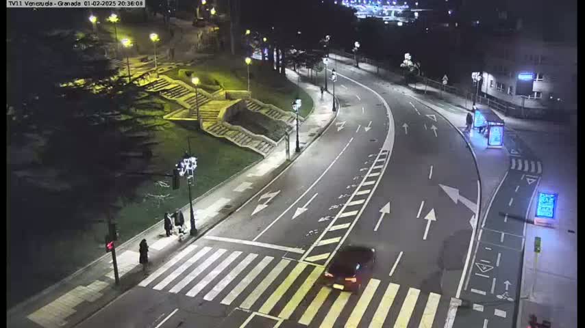 A nighttime, well-lit street scene shows a car approaching a crosswalk where pedestrians are walking,  with stairs and landscaping visible on one side of the road.