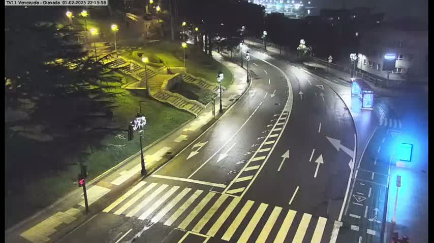 A wet, nighttime street scene shows a curving road with pedestrian crossings, streetlights, and stairs leading up to a park-like area.
