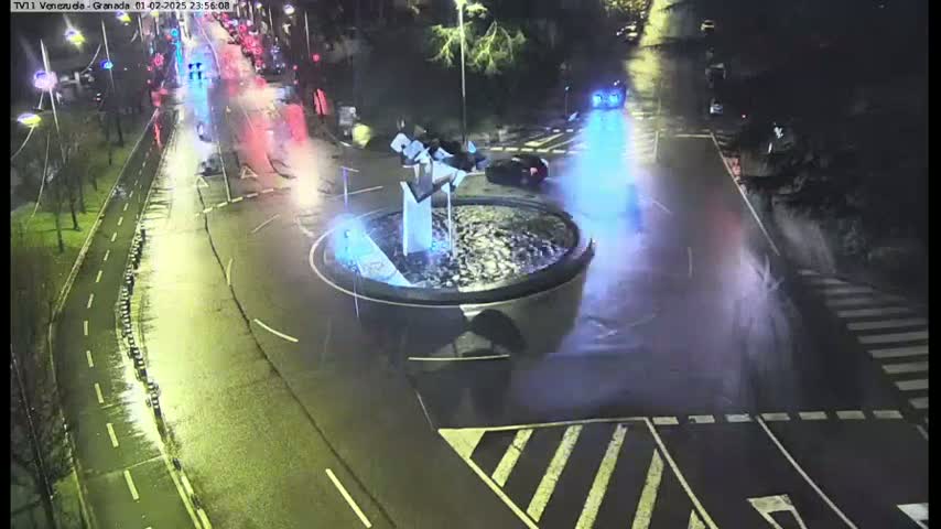 A nighttime, wet road scene shows a roundabout with a fountain in the center, surrounded by cars and pedestrian crossings.