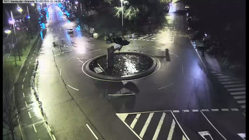 A wet, dark roadway curves around a circular traffic island with a dark sculpture at its center, illuminated by streetlights.