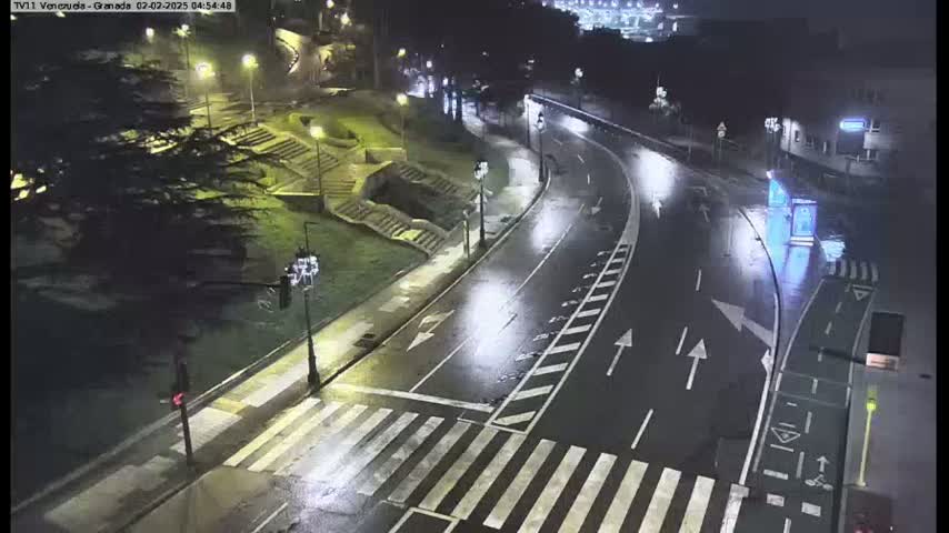 A wet, empty road curves through a nighttime city scene, illuminated by streetlights, with a pedestrian crossing and some greenery visible.
