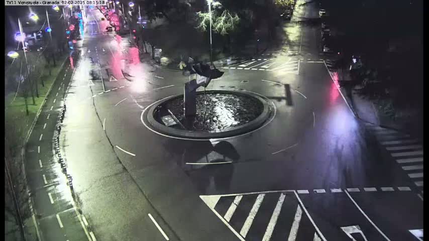 A wet, nighttime street scene shows a roundabout with a central sculpture, surrounded by roads with sparse traffic and pedestrian crossings.