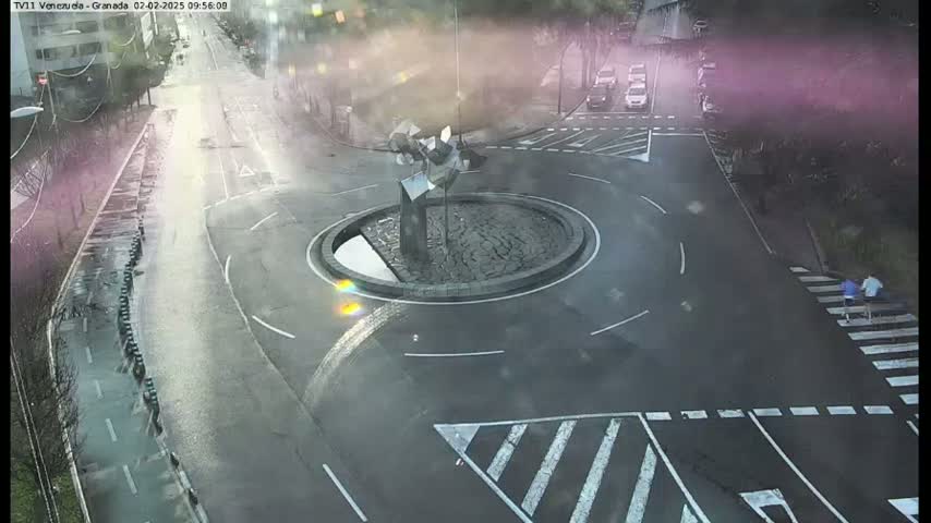 A wet roundabout with a modern sculpture in the center is surrounded by roads and crosswalks, with a few cars and pedestrians visible on a cloudy day.