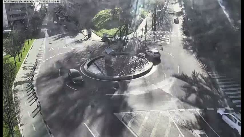 A high-angle view shows a roundabout with a central fountain, several cars, and surrounding roads and trees under a sunny sky.