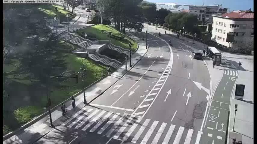 A sunny day reveals a curving road with pedestrian crossings, flanked by green landscaping and stairways, leading to buildings in the background.