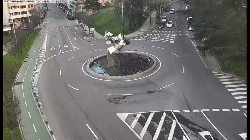 A mostly empty roundabout with a modern metal sculpture in the center, surrounded by roads with light vehicle traffic and a bike lane, on a seemingly overcast day.