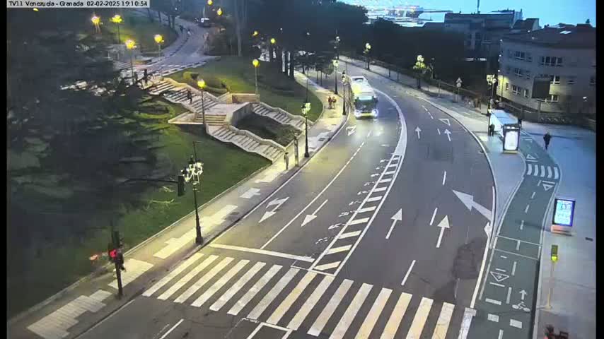 A nighttime, high-angle view shows a curved roadway with a bus and pedestrians, adjacent to a grassy area with stairs and trees.