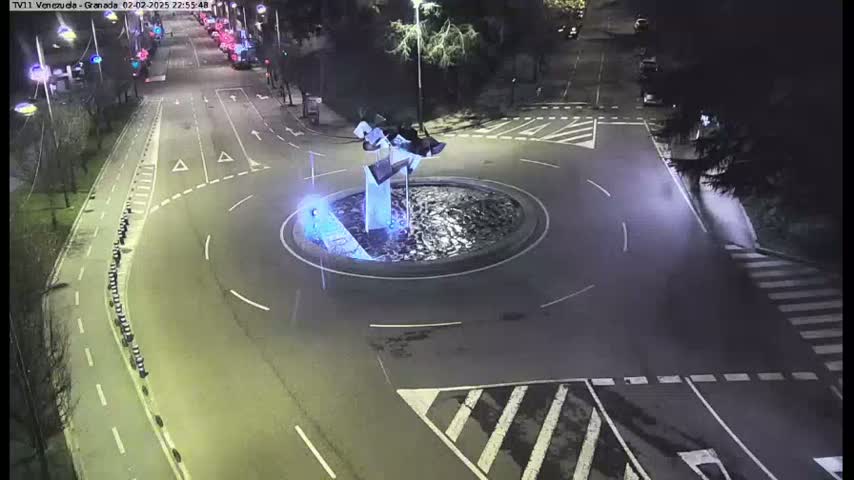 A nighttime aerial view shows a roundabout with a modern sculpture in the center, surrounded by roads with sparse traffic.