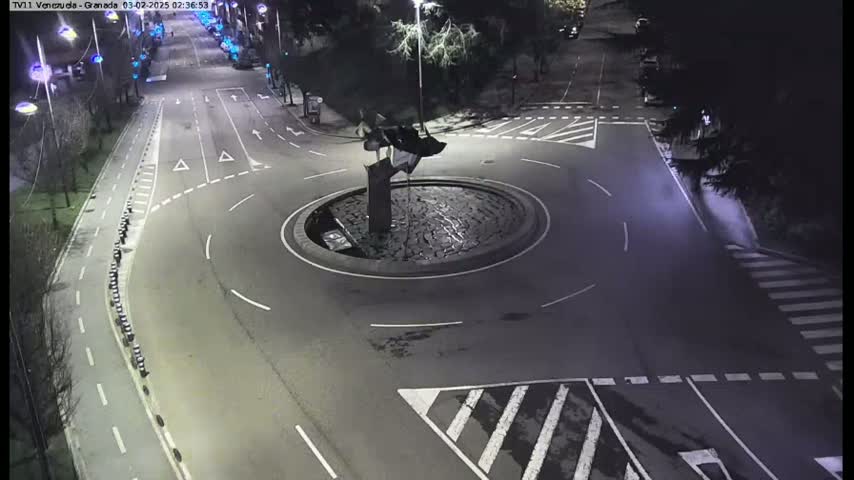 A nighttime aerial view shows a traffic circle with a dark, abstract sculpture in the center, surrounded by roadways with white lane markings and pedestrian crosswalks.