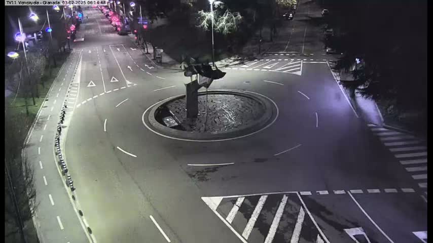 An empty roundabout at night, surrounded by roadways and pedestrian crossings, with a dark, indistinct sculpture at its center.