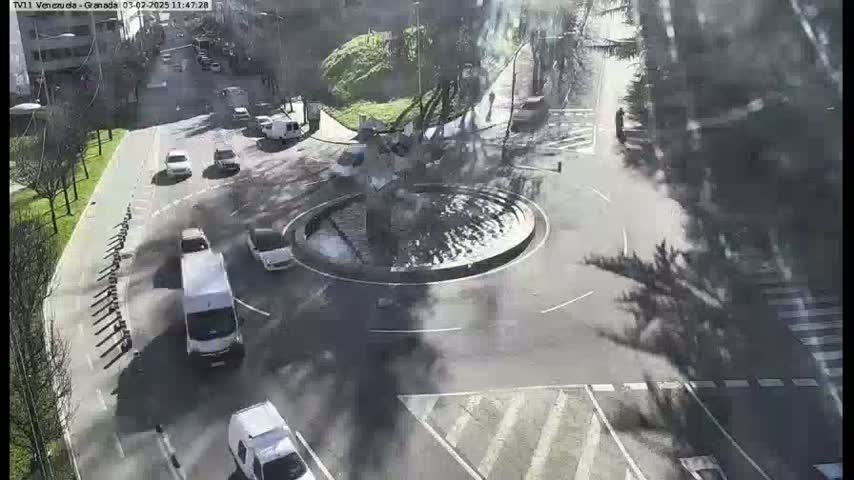Several cars are driving around a traffic circle on a sunny day.