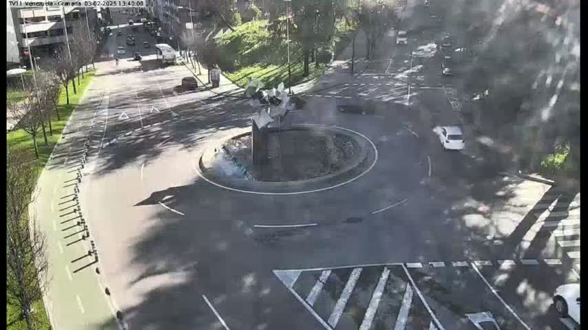A sunny overhead view shows a roundabout with a modern sculpture in the center, surrounded by roadways, bike lanes, and some parked cars.