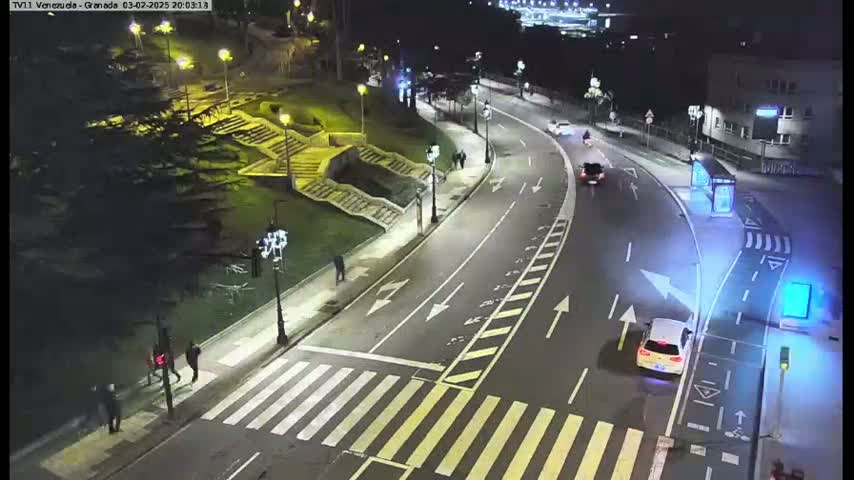 A nighttime, brightly lit street scene shows a curving road with crosswalks, pedestrians, and several vehicles, including a white car near the bottom right.