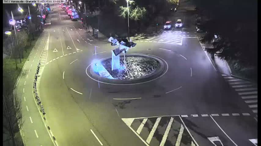 A nighttime aerial view shows a traffic circle with a modern sculpture in the center, surrounded by roads and crosswalks.
