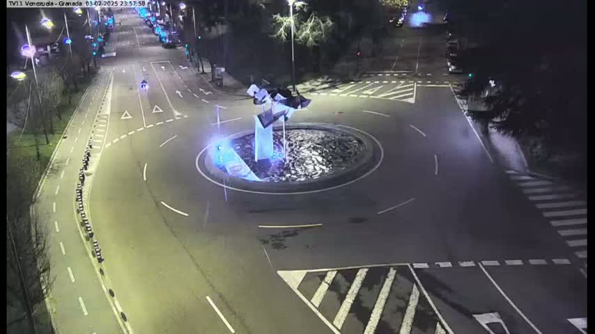A nighttime aerial view shows a roundabout with a modern sculpture in the center, surrounded by roads and sparsely parked cars.