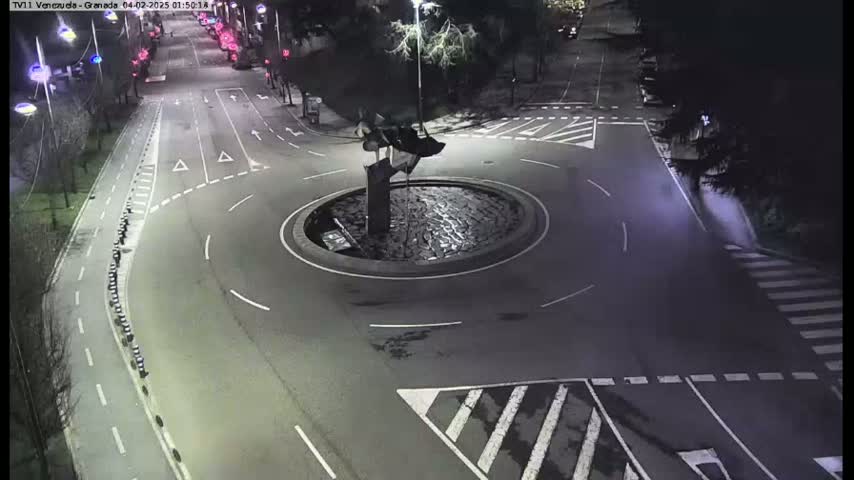 A nighttime aerial view shows a roundabout with a dark sculpture in the center, surrounded by roads with light-colored markings under artificial light.