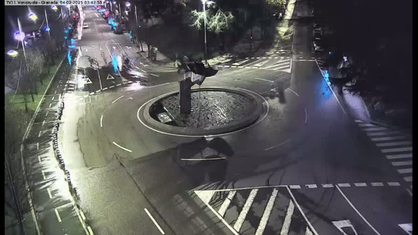A nighttime, wet roadway intersection features a circular traffic island with a fountain in the center.