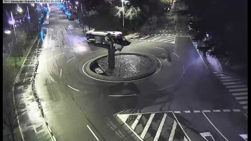 A wet, nighttime view of a traffic circle with a bus parked near its center, surrounded by roads and street markings.
