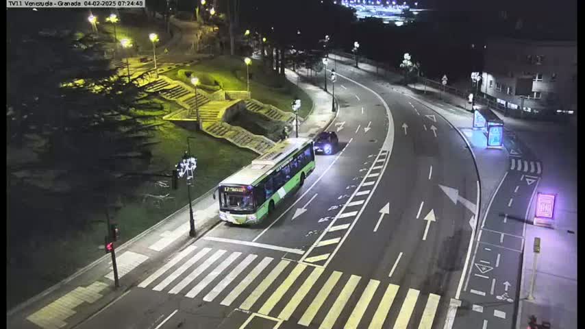 A green bus and a car are stopped at a crosswalk on a curved road at night.