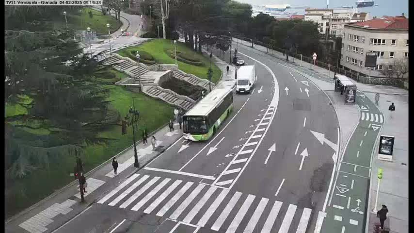 A city street scene on an overcast day shows a bus and van stopped in traffic near a pedestrian crossing and bike lane, with a staircase and green space visible on one side.