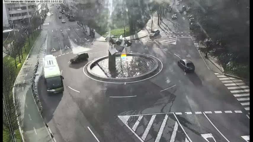 A wet, overcast city intersection shows a roundabout with a sculpture in the center, surrounded by several cars and a bus.