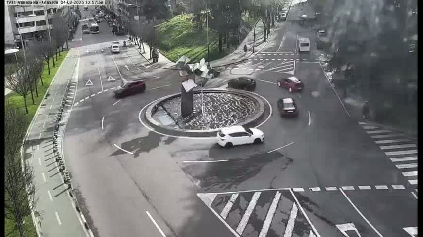 A wet roundabout with several cars and a central fountain is surrounded by roads and pedestrian crossings.